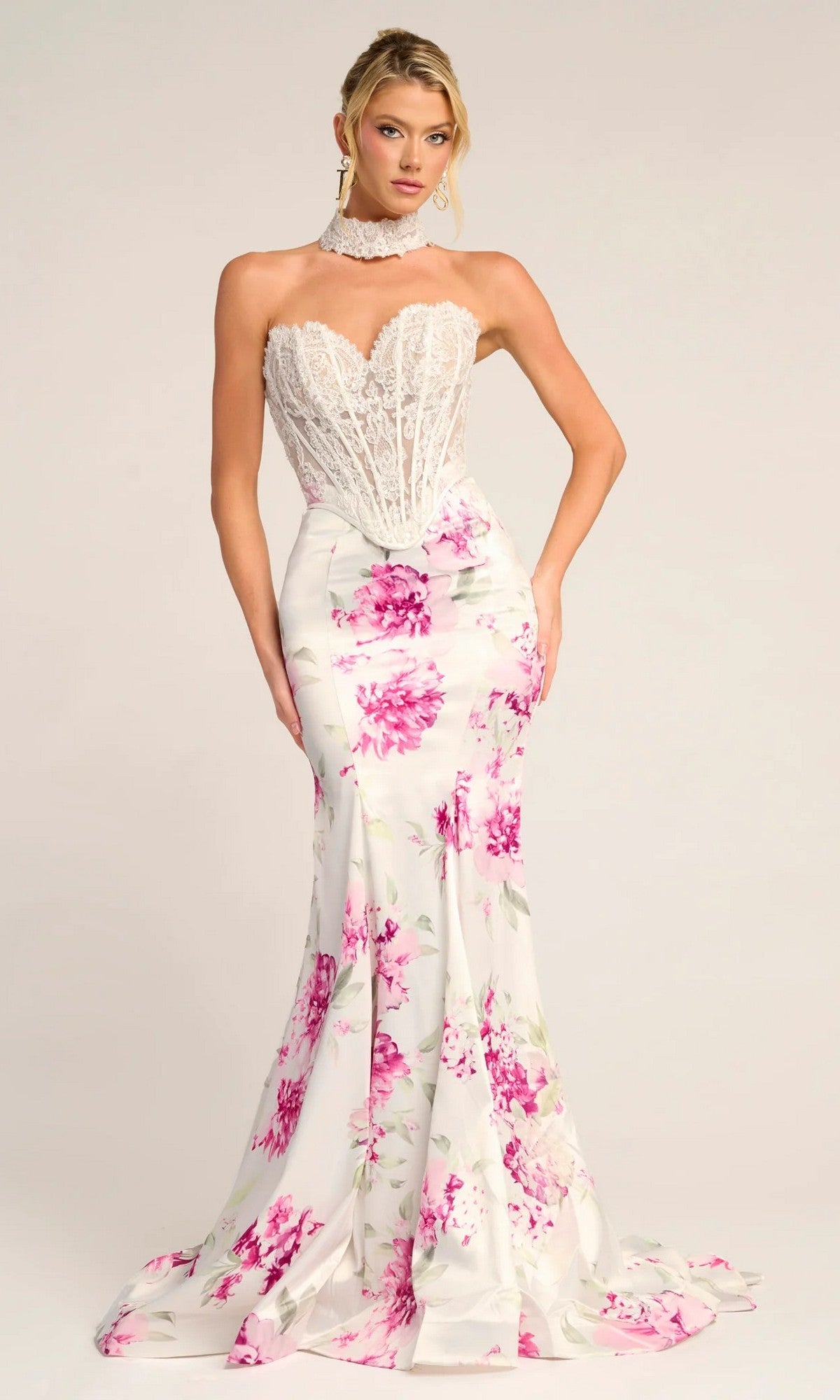 A woman wears the Long Formal Dress: Portia and Scarlett PS26299 in White/Pink, featuring a strapless lace corset bodice and a fitted satin formal skirt with pink floral details.